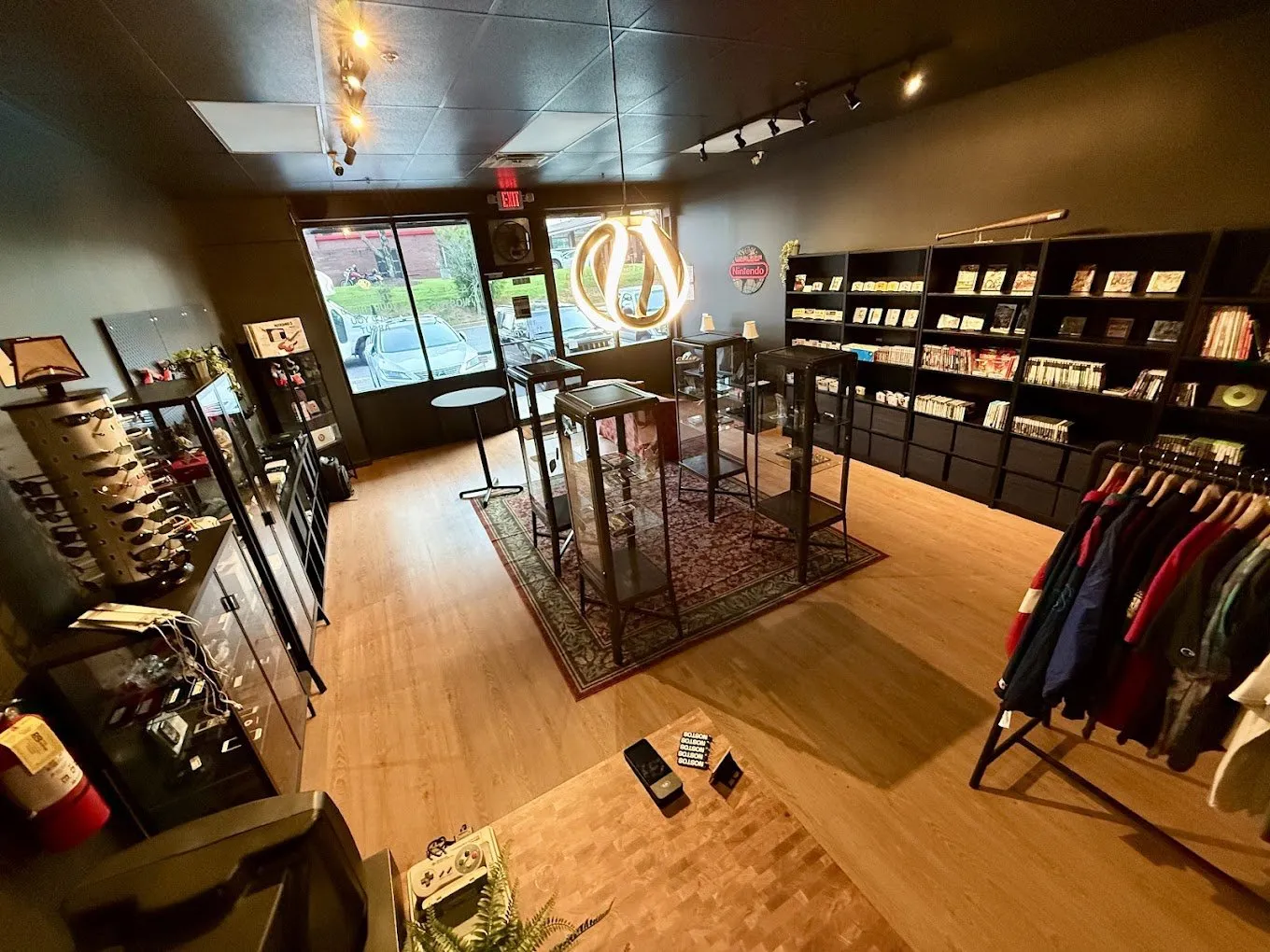 Inside the NOSTOS storefront in Duluth, Georgia: lit glass display cases of games and handhelds on a patterned rug, black wall shelves of boxed titles, a rack of vintage apparel, and the gold ring logo glowing over the entrance.